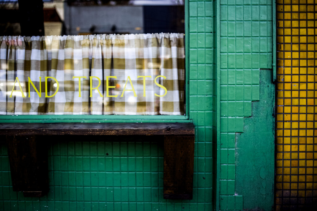 Tiled storefront and window with gingham curtains and yellow “AND TREATS” lettering.