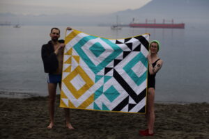 Two people in swim suits, standing on a misty winter beach, hold up a quilt.
