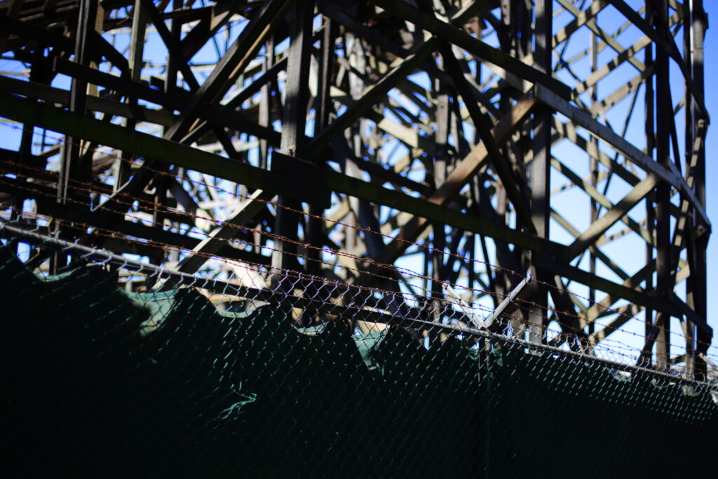 An old, wooden roller coaster track, behind a chain-link fence.