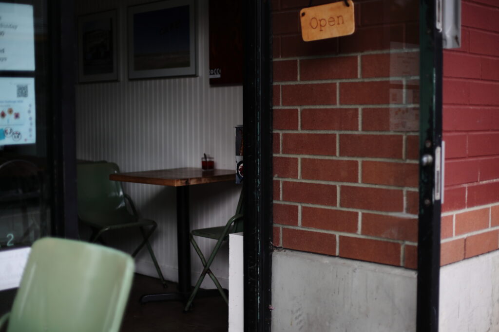 An open glass door reveals the inside of a small cafe with green chairs sitting empty at a wood table.