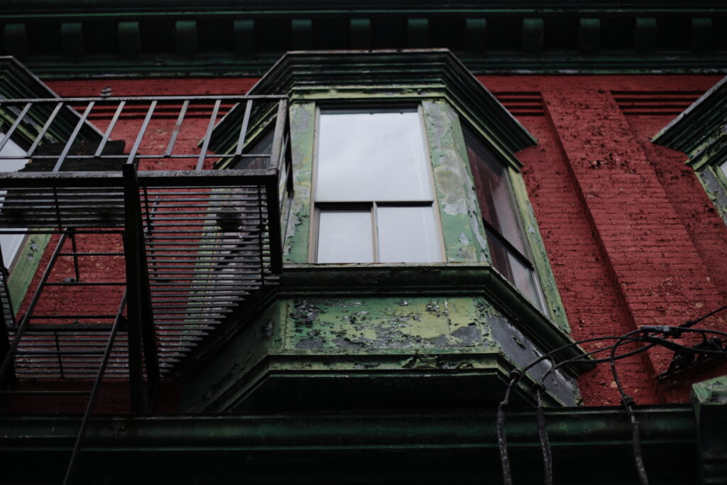 A dilapidated Edwardian bay window juts out from a brick wall with peeling red paint.