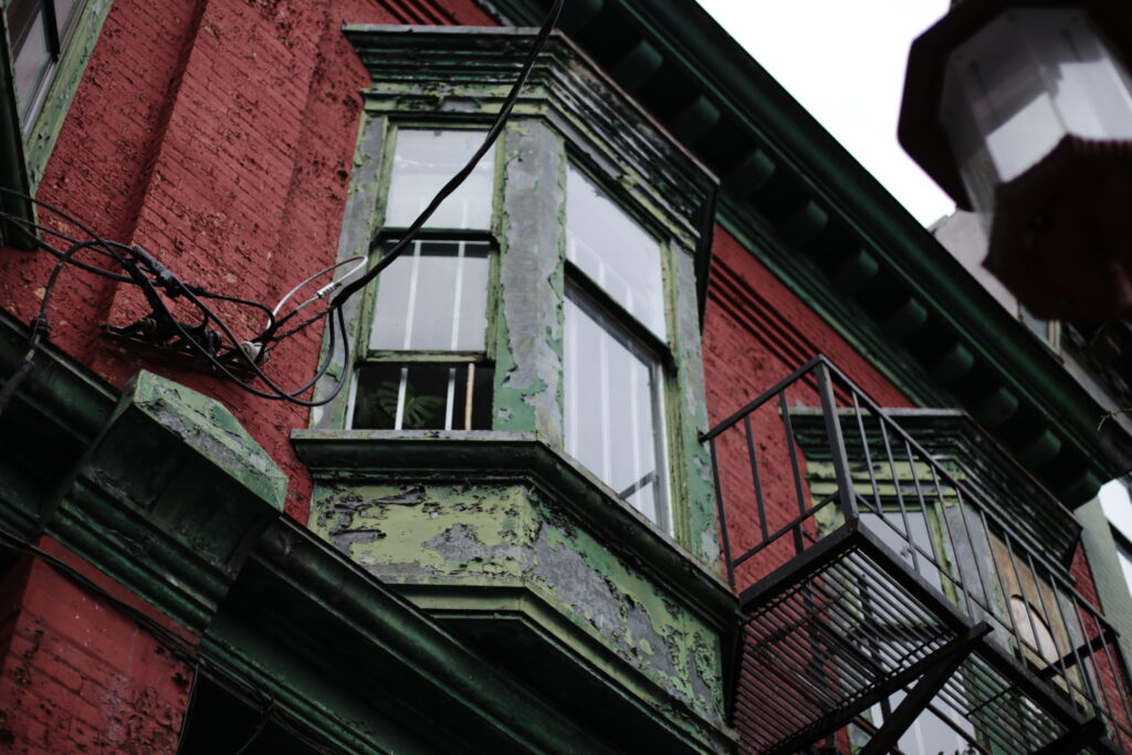 A dilapidated Edwardian bay window with a wrought iron fire escape juts out from a brick wall with peeling red paint.